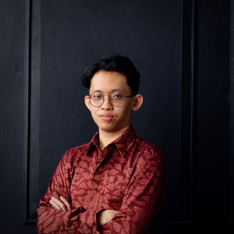Portrait of Muhammad Nuril Huda wearing a maroon batik shirt against a dark background